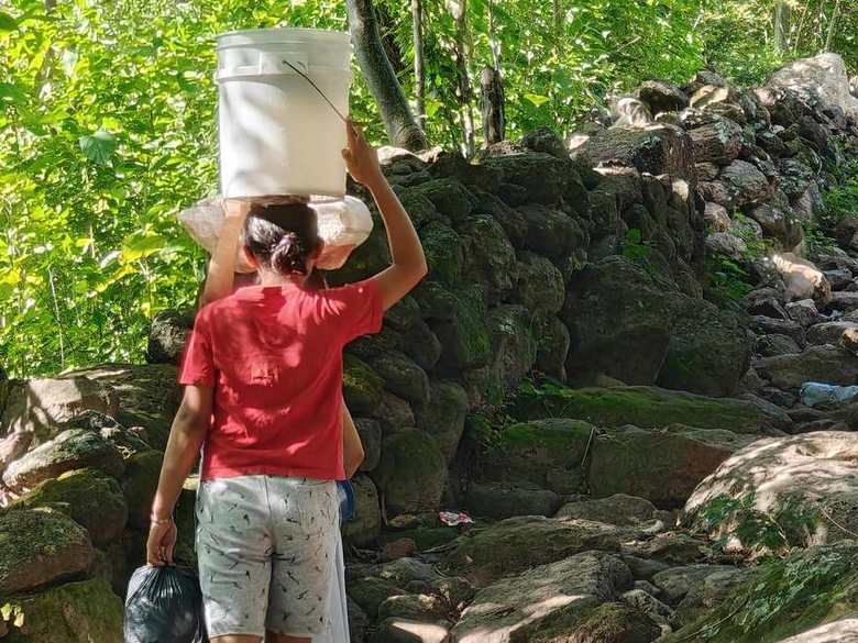 A girl carries a large white bucket of water on her head up a rocky path in El Guanito